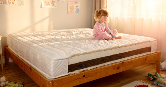 Enfant assis au bord d’un grand matelas blanc sur lit en bois dans une chambre lumineuse, décorée de dessins muraux.