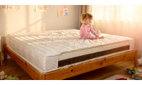 Enfant assis au bord d’un grand matelas blanc sur lit en bois dans une chambre lumineuse, décorée de dessins muraux.